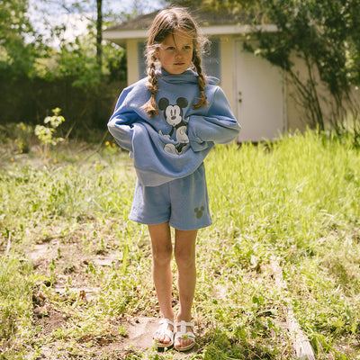 Child standing in a park wearing a cute kid's Disney outfit—blue Mickey Mouse matching set with kid's Disney sweatshirt and kid's Disney shorts. 