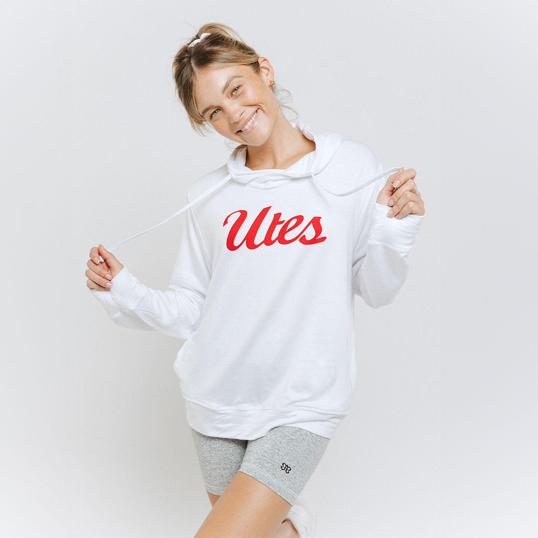 A woman wearing a white University of Utah hoodie with the logo 'Utes' printed in red on the front, paired with grey shorts.