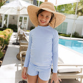 Child wearing a blue striped children's rash guard and wide-brimmed hat by a poolside.