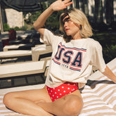 Woman wearing a 'USA 1776' t-shirt and red swimsuit with white stars, sitting on a sunbed outdoors.