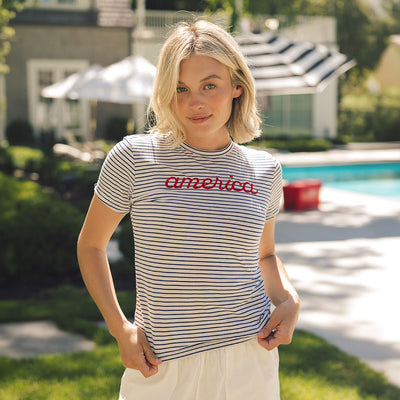 Woman wearing a striped t-shirt with 'america' printed on it, standing outdoors near a pool.