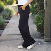 A person wearing black wide-leg drawstring pants with side pockets, paired with a white top and white sneakers, standing on a stone pathway in a garden setting.