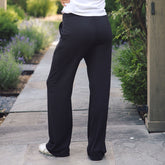 A person wearing black wide-leg pants and a white top, standing on a stone walkway outdoors with greenery and a wooden post in the background.