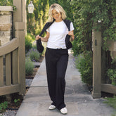 A woman walking outdoors on a stone path between wooden gates, wearing a white crew-neck T-shirt, black wide-leg pants, a black open-front cardigan, and white sneakers.