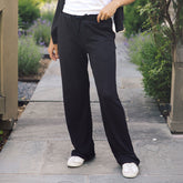 A person wearing black wide-leg pants, a white top, and white sneakers standing on a stone pathway outdoors with greenery on both sides.