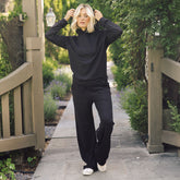 A woman stands on a stone walkway outdoors wearing a black long-sleeve mock-neck top and matching black wide-leg pants, with white sneakers. Trees, shrubs, and wooden gateposts are visible in the background.