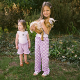 Two young girls in a garden holding a baby goat wearing cute kid's pants for Disneyland in purple check Minnie Mouse print.