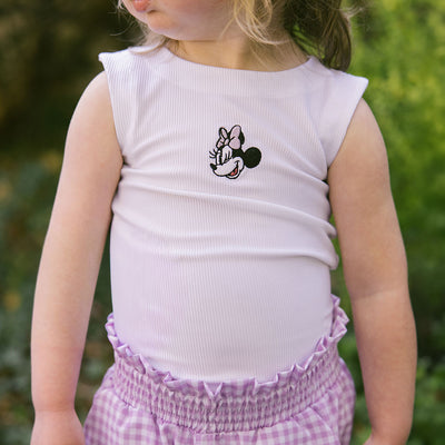 Close up of child wearing a Minnie Mouse tank top for kids with purple gingham Disney shorts for kids on a blurred green background.