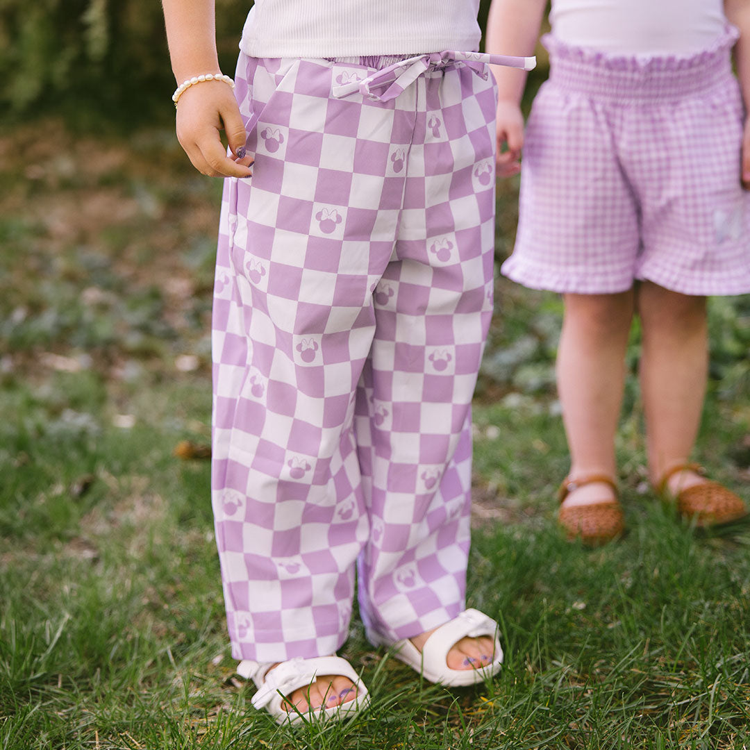 Two children wearing purple checkered pants with Minnie Mouse print standing on grass.