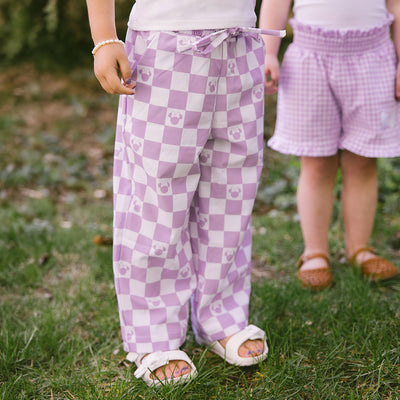 Two children wearing purple checkered pants with Minnie Mouse print standing on grass.