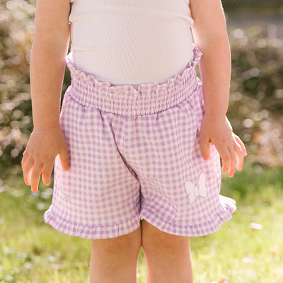 Close up of child in a garden wearing purple gingham kid shorts for Disneyland with Minnie Mouse embroidered bow on the leg. 