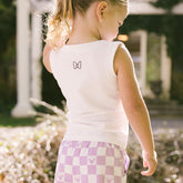 Child wearing a white sleeveless shirt with a Minnie Mouse bow design and purple checkered pants in an outdoor setting.