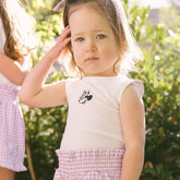 Young girl wearing a Minnie Mouse tank top with purple gingham shorts on a blurred natural background.