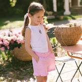 Young girl in a white sleeveless top and pink checkered shorts—cute Disney themed outfit for girls—standing outdoors with flowers in the background