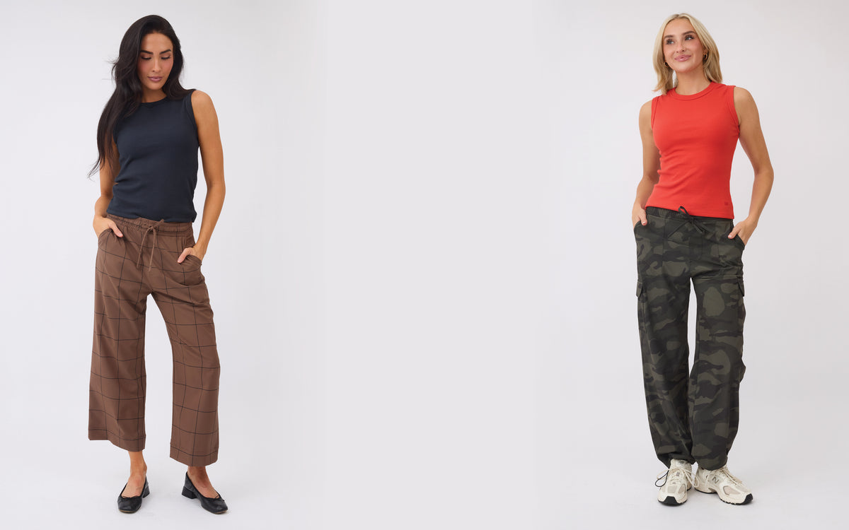 Two women modeling sleeveless tops and wide-leg cargo pants against a plain light gray studio background. The left model wears a dark navy sleeveless top with brown plaid pants, and the right model wears a bright red sleeveless top with dark camouflage cargo pants.