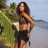 Woman in a bikini and sarong standing on a beach with palm trees and ocean in the background