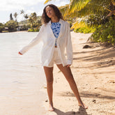 Woman in white cover-up shirt and shorts over a blue floral swim top standing on a sandy beach with palm trees in the background