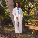 Woman in a white linen pants and button down shirt over a swimsuit standing under a palm tree in a tropical setting
