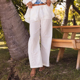 Woman wearing white beach pants standing next to a wooden picnic table in a park setting.