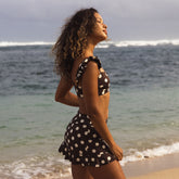 Woman in a brown and white polka dot swimsuit standing on a beach with ocean waves in the background.