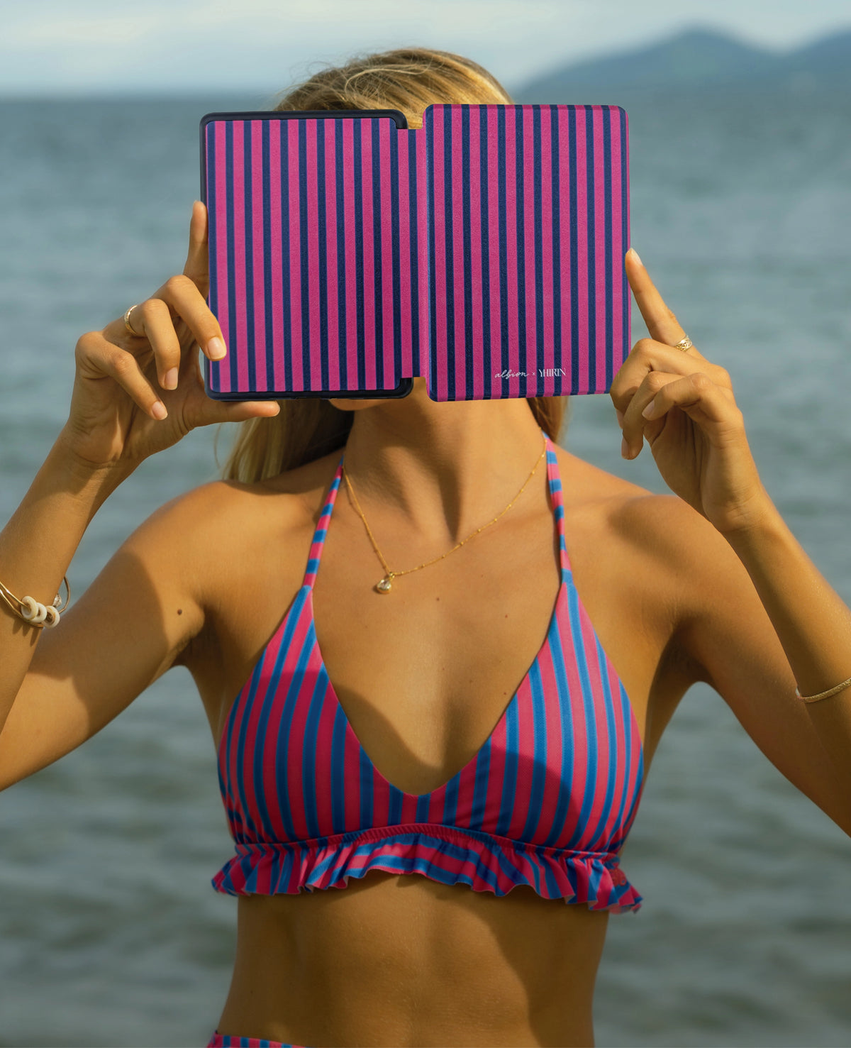 Person wearing a striped swimsuit holding a pink and blue striped product in front of their face with water and mountains in the background.