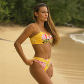 Woman in a yellow floral bikini standing on a sandy beach with greenery in the background