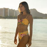 Woman in a yellow floral bikini standing by water with buildings and mountains in the background