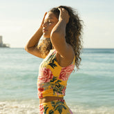 Woman in a floral yellow bikini top standing on a beach with ocean in the background
