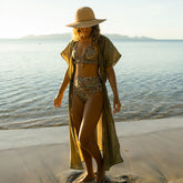 Woman in a zebra bikini with a green wrap cover up and sun hat walking on a beach with ocean in the background