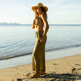 Woman in a sun hat and green coverup pants over a two piece swimsuit standing on a beach with ocean in the background
