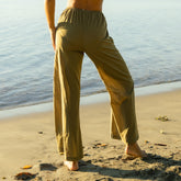 Woman wearing olive green cover up pants standing on a beach with water in the background