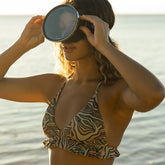 Woman in a neutral zebra print bikini  with ruffles standing by the water, holding goggles to her eyes.