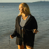 Woman standing by the ocean wearing a striped bikini and a black oversized swimsuit cover-up shirt