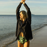 Woman stretching her arms above her head on a beach, wearing a black and white bikini top, palm green high waisted bottoms, and a black wrap cover up top.