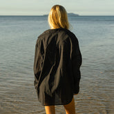 Woman wearing a black oversized collard shirt standing on a beach with water in the background