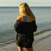 Back view of woman wearing an oversized black shirt over a swimsuit standing on a beach looking out at the ocean