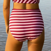 Back view of woman wearing high waisted, full coverage bikini bottoms in pink stripe print with blurred ocean background.