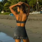 Woman in a black bikini top and skirt standing on a beach with boats and greenery in the background.