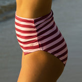 Side view of woman wearing flattering ruched bikini bottoms with a mid rise and the ocean blurred in the background. 