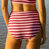 Back view of woman wearing full coverage bikini bottoms with mid rise and flattering ruched side on a blurred ocean background.