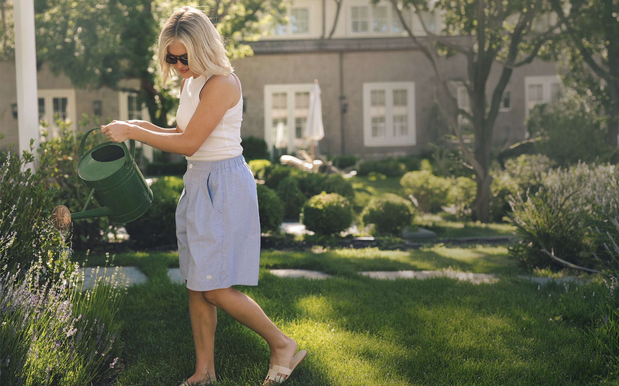 A woman in a white tank top and light blue shorts watering a garden bed with a green metal watering can in a sunny, landscaped backyard.