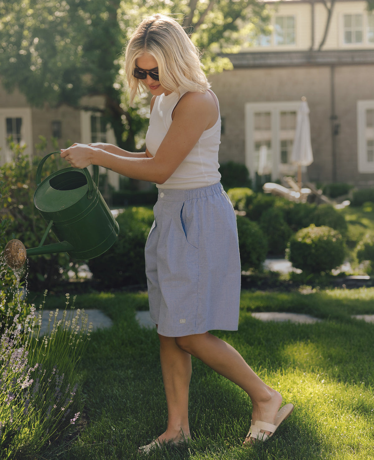 A woman in a white tank top and light blue shorts watering a garden bed with a green metal watering can in a sunny, landscaped backyard.