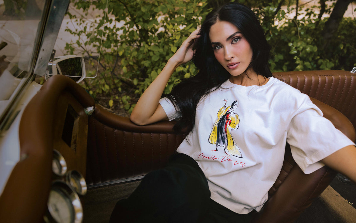 A woman with long dark hair sits in a brown leather car interior, wearing a white oversized T-shirt with a colorful graphic of a woman in a yellow dress and the text 'Diva for Life' printed below it.