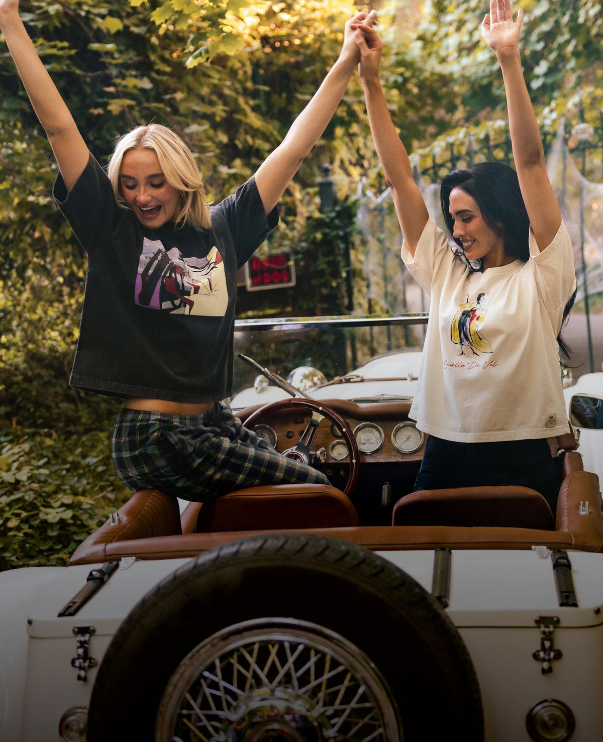 Two women sitting in the back of a vintage convertible car, holding hands with arms raised. The woman on the left wears a black graphic t-shirt and plaid pants, while the woman on the right wears a cream graphic t-shirt and dark bottoms.