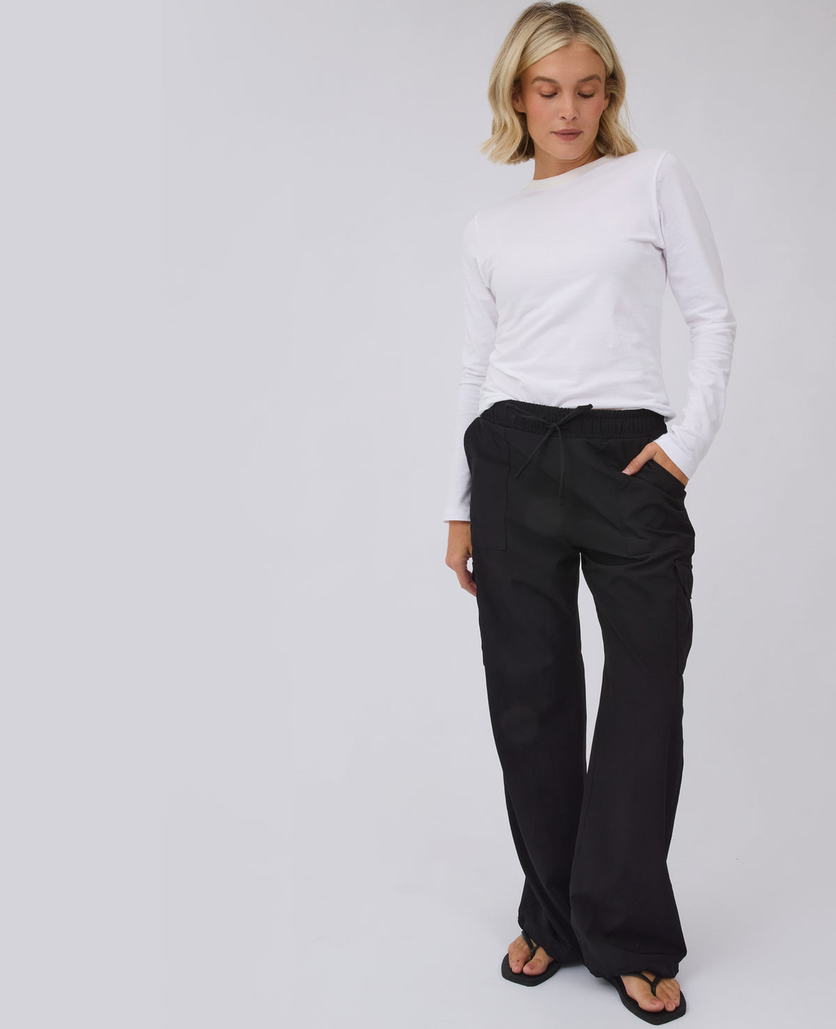 A woman modeling a white long-sleeve top and black wide-leg trousers against a light gray studio background.