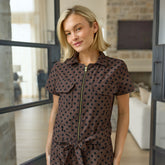 Close up of woman wearing a short-sleeved jumpsuit in brown and black polka dot print with gold zipper details standing against large glass doors.