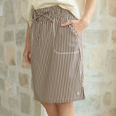 Woman wearing a brown striped knee length skirt with pockets and adjustable front tie in front of a blurred stone wall.