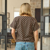 Back view of woman wearing a brown polka dot shirt and blue jeans standing in a room with a stone wall.