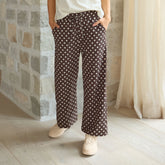 Woman wearing brown polka dot, wide leg pants with sneakers standing against a stone wall. 