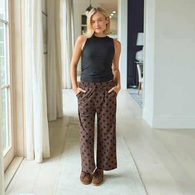 Woman wearing a black sleeveless top and brown polka dot pants standing in a well-lit room.